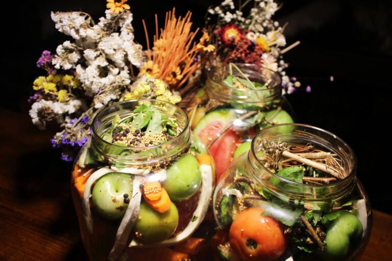 Three glass jars of pickled vegetables: green and red tomatoes, carrots, mustard seeds and herbs, in front of a bunch of dried flowers